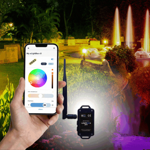 A hand holds a smartphone with a color wheel lighting app, connected to the Outdoor RGBW 4-Ch Wi-Fi LED Controller ~ wLightBox Pro by BleBox. In the background, garden fountains glow in vibrant colors at night.