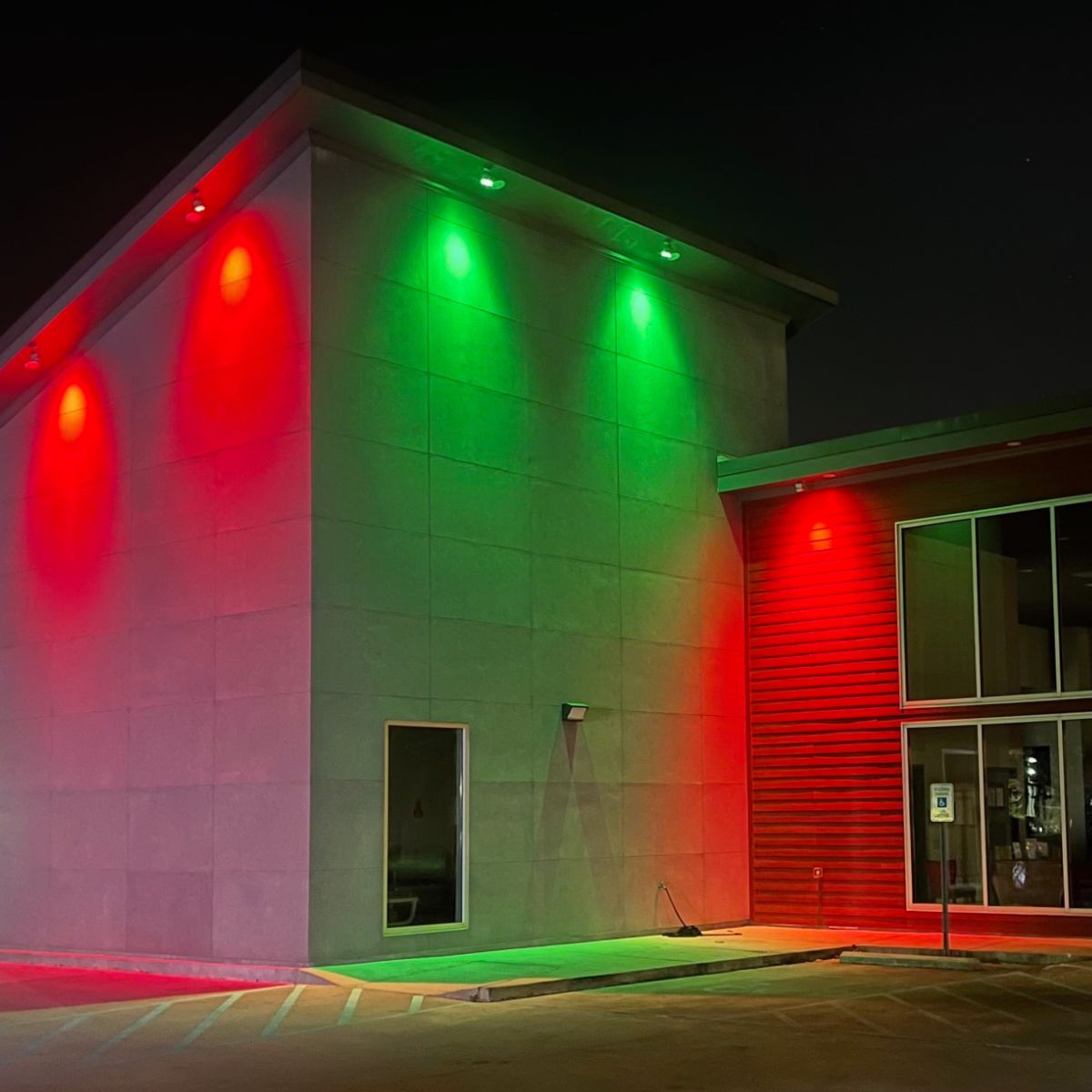 A modern building at night glows with green and red Revo ColorFlow Casambi Series color-changing outdoor LED wall washer lights, casting vibrant reflections on walls and ground. The empty parking lot features a visible handicap parking sign near the entrance.