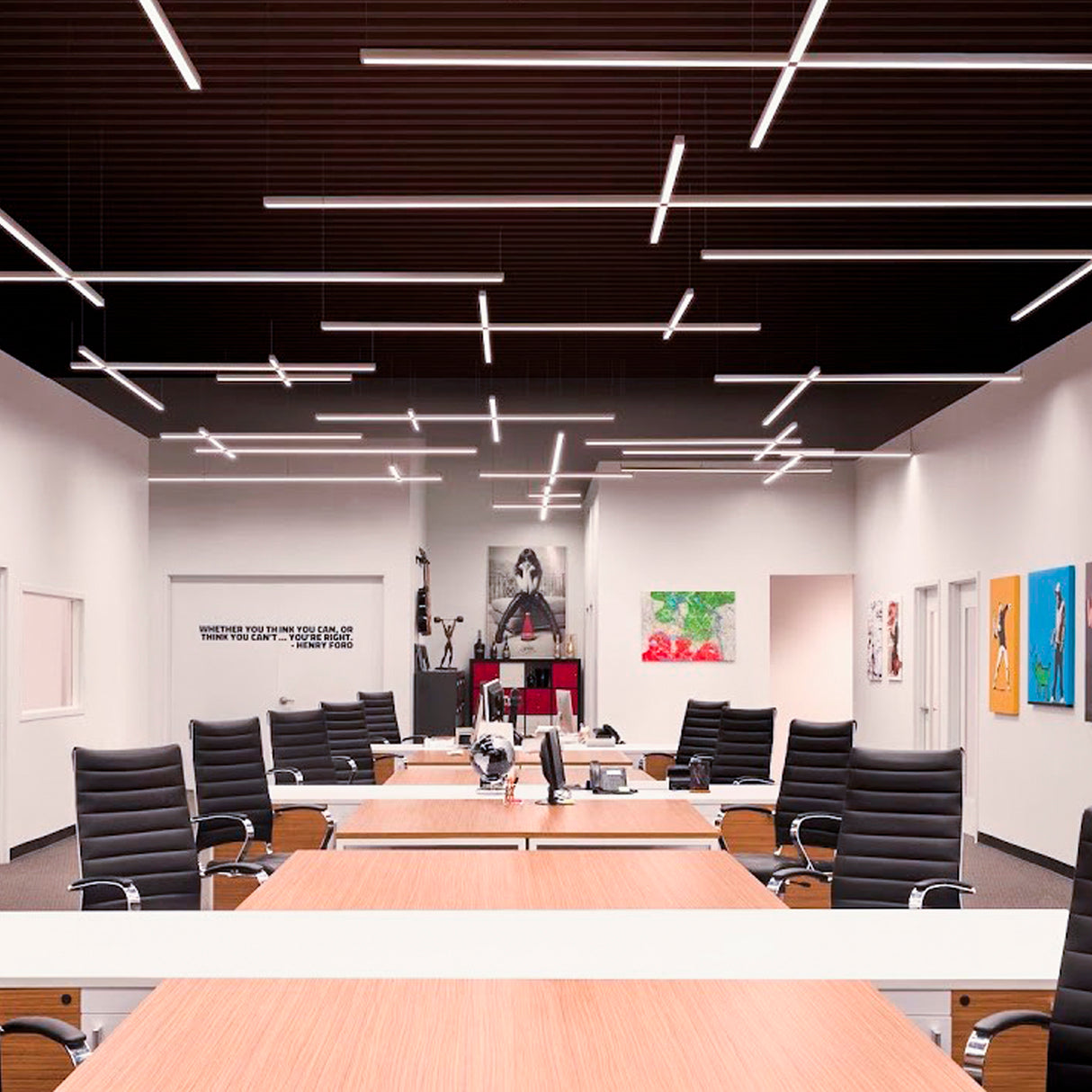 A modern office with long wooden desks, black swivel chairs, and computers is illuminated by the Waterproof LED Channel ~ Model Roma Easy On IP65. Minimal décor and colorful artwork decorate the white walls.