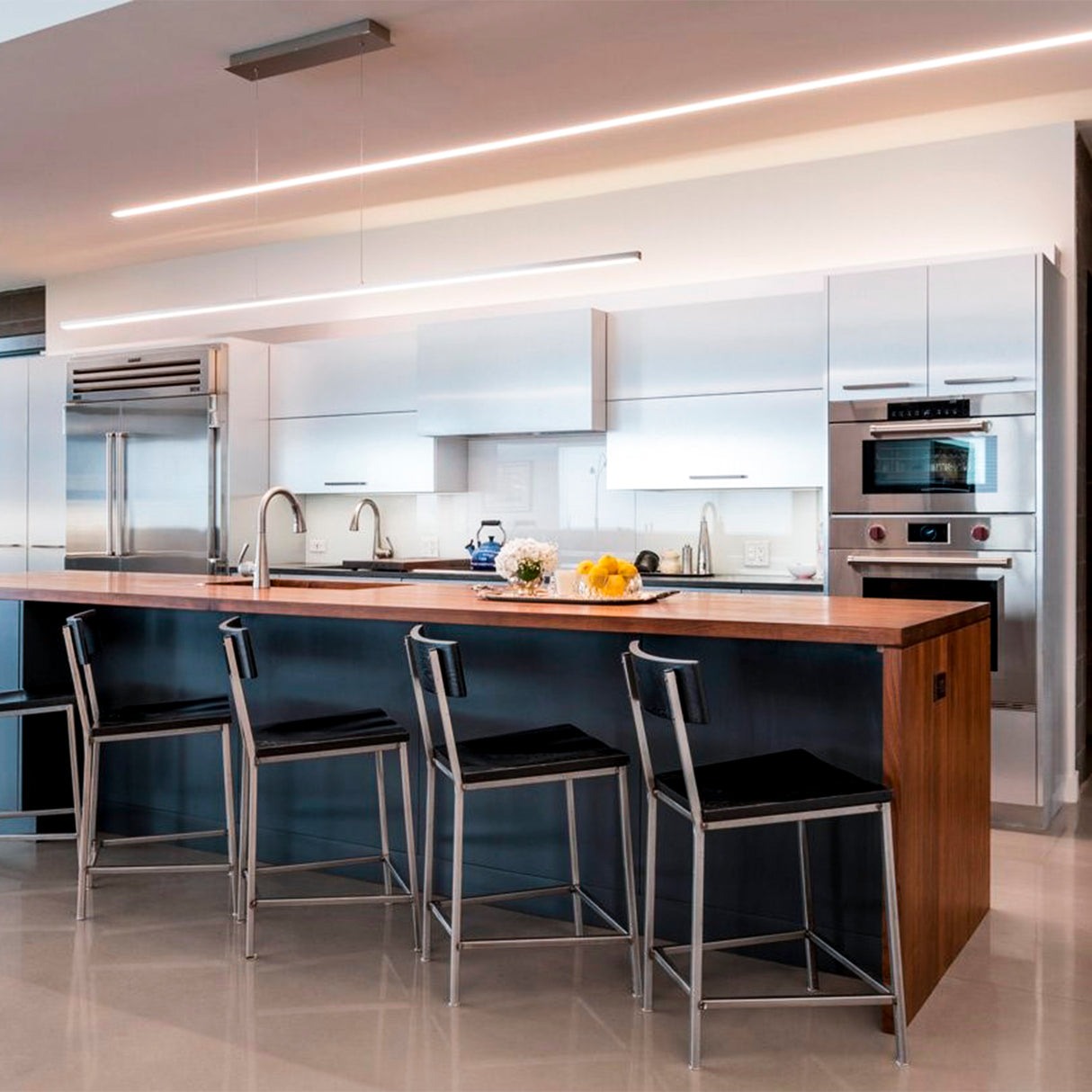 A modern kitchen with a large island, three metal bar stools, stainless steel appliances, and white cabinets. The Waterproof LED Channel ~ Model Roma Easy On IP65 adds a sleek touch above the wooden countertop with flowers and fruit.