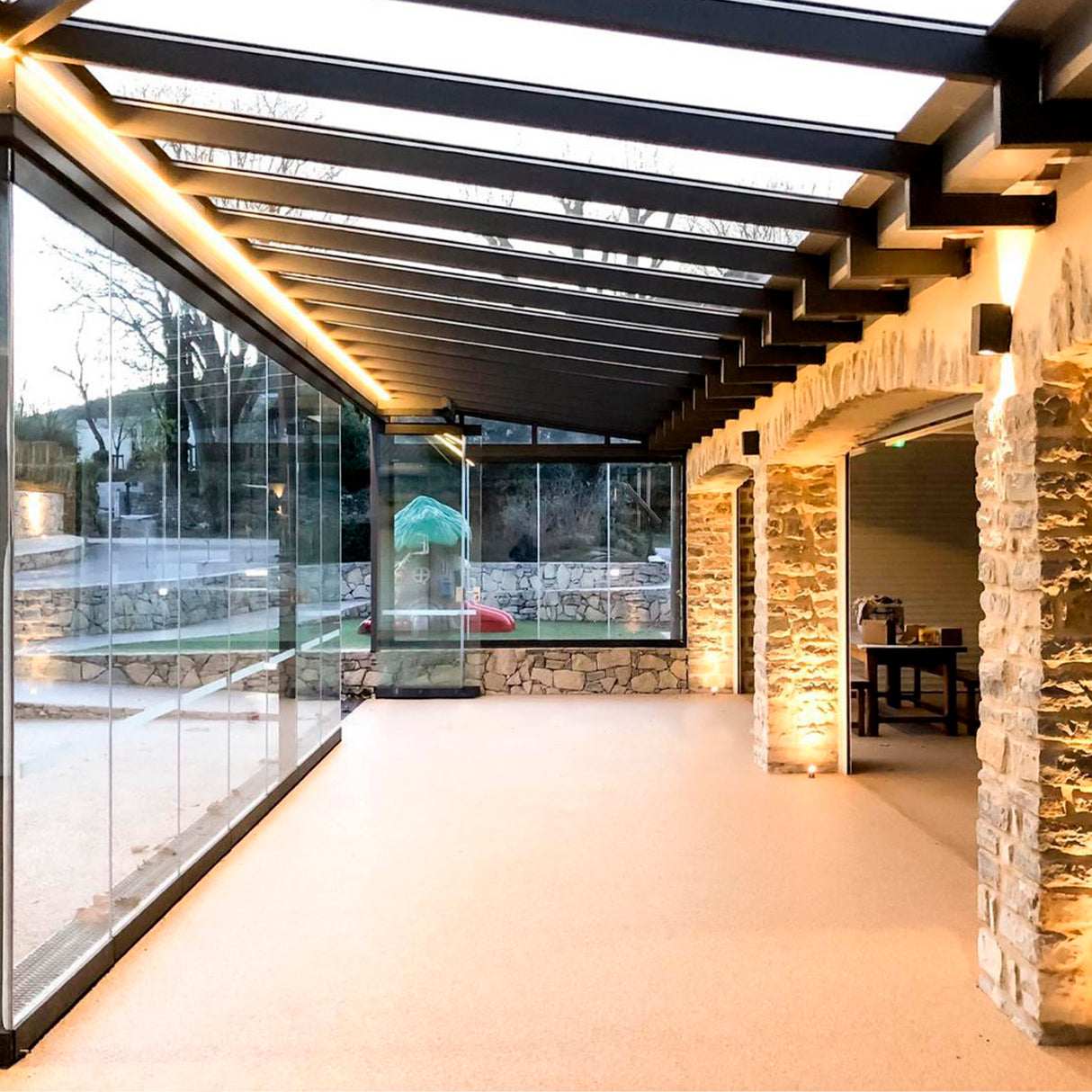 A modern glass-walled room with a stone accent wall, exposed ceiling beams, and warm lighting from the Waterproof LED Channel ~ Model Roma Easy On IP65, overlooking trees and a small green playground visible outdoors.