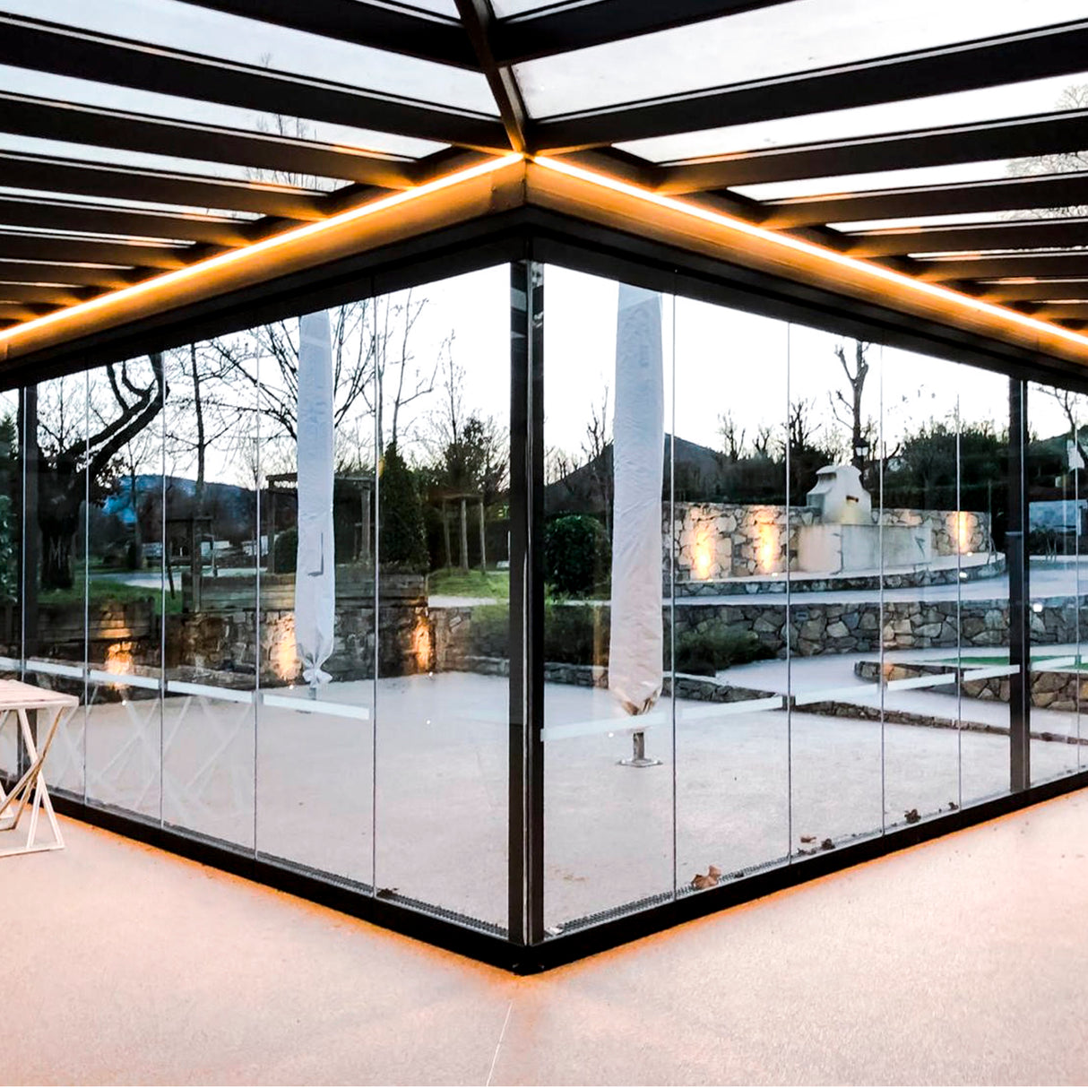 A modern glass-walled patio with a black metal frame and warm yellow ceiling lighting uses the Waterproof LED Channel ~ Model Roma Easy On IP65. The outdoor area features stone walls, trees, and ambient waterproof lighting.