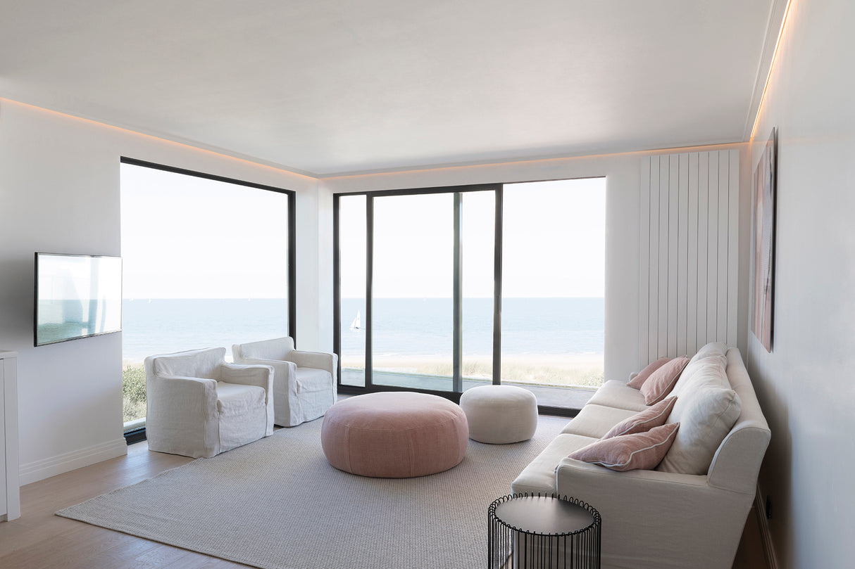 Minimalist living room with white walls, large window, and sliding glass doors to the ocean. Waterproof LED Channel ~ Model Roma Easy On IP65 highlights the white sofa, two chairs, pink ottoman, and light rug for a sleek modern vibe.