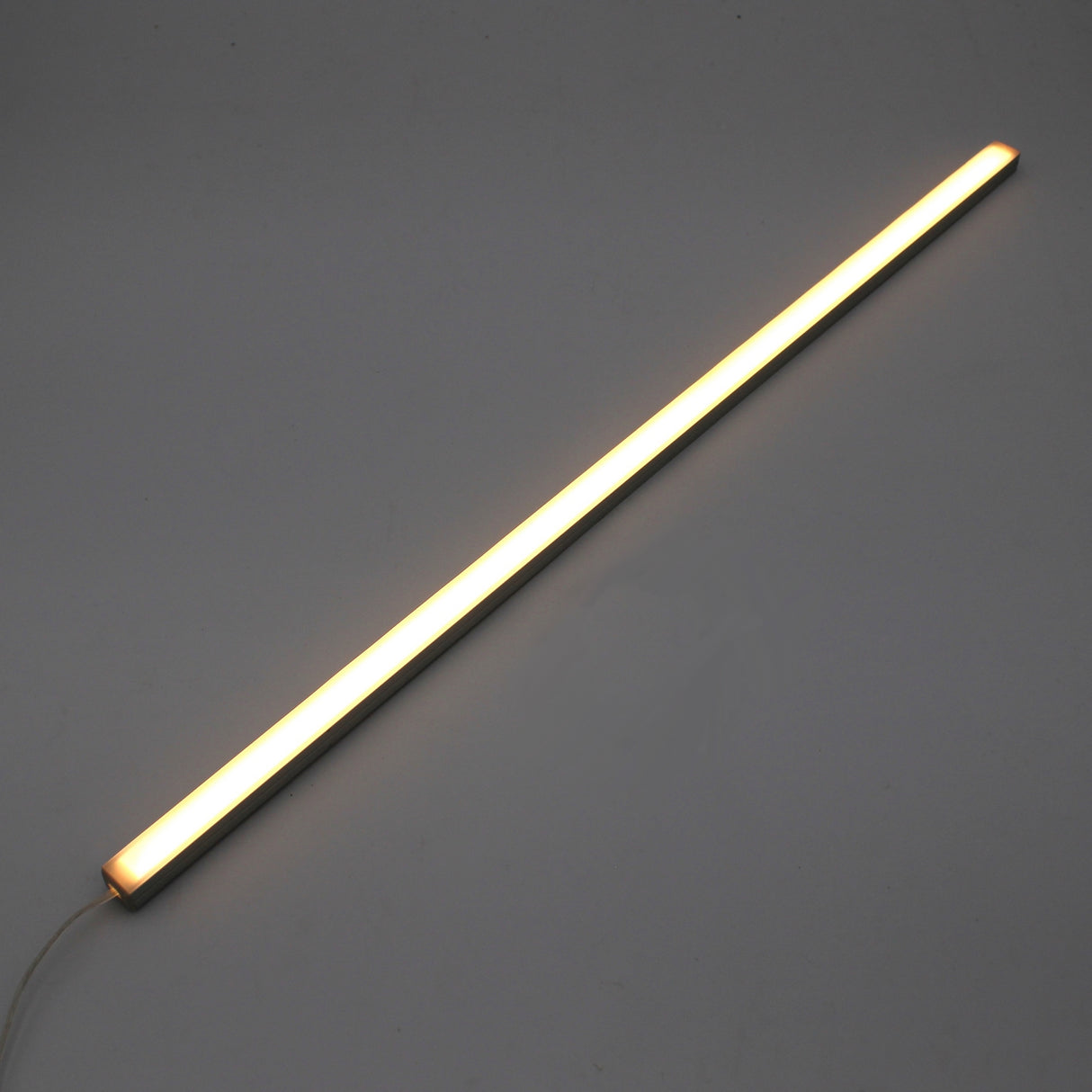 The White Universal Surface-mount Waterproof Outdoor LED Lighting Fixture (24V) ~ Roma Series emits a warm white glow as a slim, dotless LED bar shown diagonally on a plain gray background, with its power cord visible for versatile outdoor use.