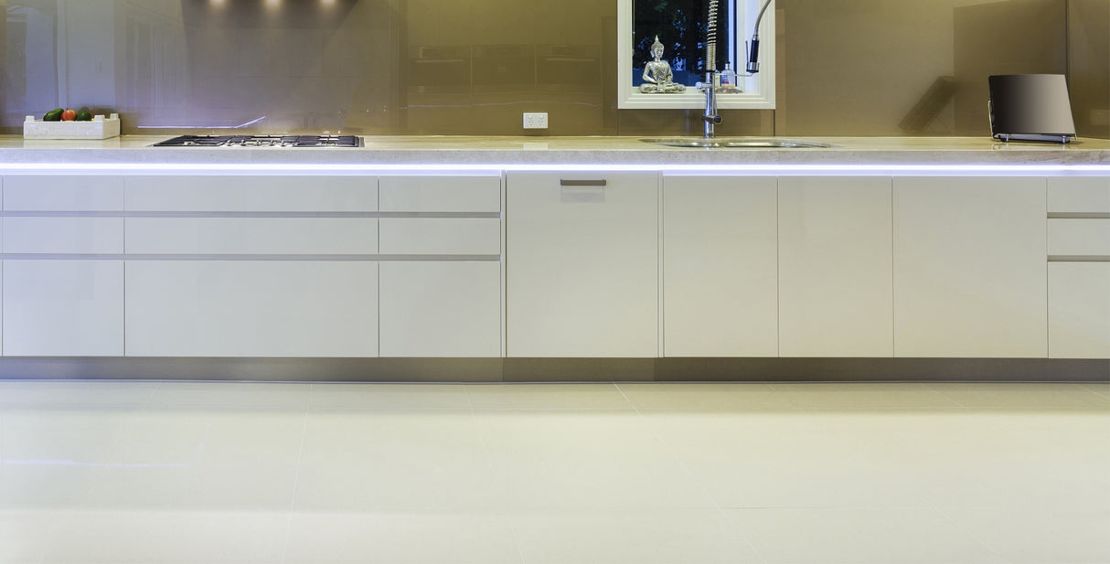 Modern kitchen with glossy white cabinets, Smart10 Miniature Surface Mount LED channel under-cabinet lighting, gas cooktop on the left, sink beneath a window in the center, beige tile flooring, and vegetables with a small appliance on the countertop.