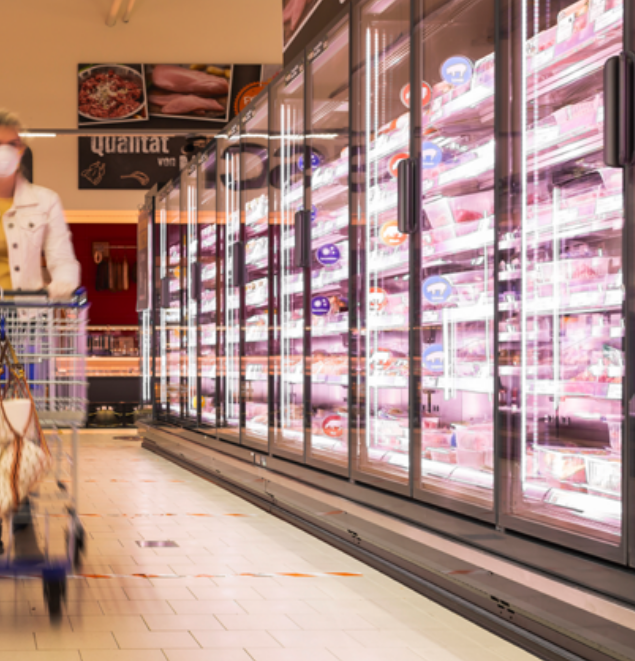 A masked shopper pushes a cart through a well-lit supermarket aisle, where glass-door freezers on the right are illuminated by the Commercial Refrigerator Lighting LED System ~ FLX Stix CDV Series.
