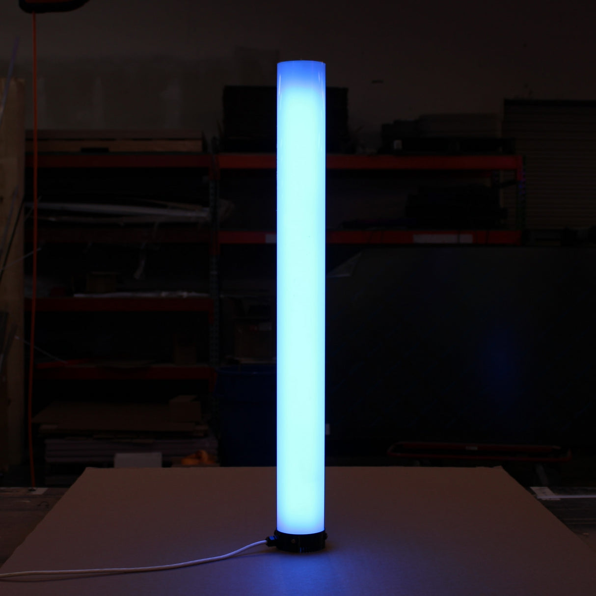 A tall Smokies100 diffuser with a Plastic Base stands vertically on a flat surface in a dimly lit room, emitting a bright blue glow, with shelves and equipment in the background.