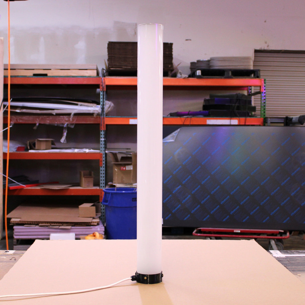 A Plastic Base for the Smokies100 Profile supports a tall, cylindrical white light fixture on a beige surface in a workshop, surrounded by shelves filled with tools and materials. An electrical cord extends from the base of the light.