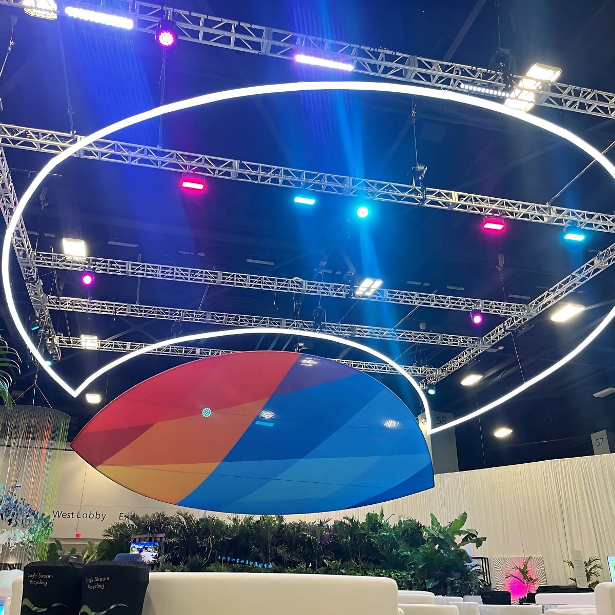 A spacious indoor event area features a suspended, multicolored 4 Acrylic Tube for LED Lights (Model Smokies100) beneath circular LEDs, with greenery and seating below. Bright, colorful spotlights highlight the vibrant scene.