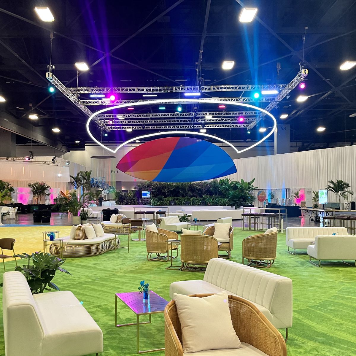 Spacious lounge with modern white sofas, wicker chairs, and small tables on a green carpet. A colorful geometric canopy and Smokies100 4 Acrylic Tube for LED Lights add ambiance from the ceiling. Lush plants decorate the area.