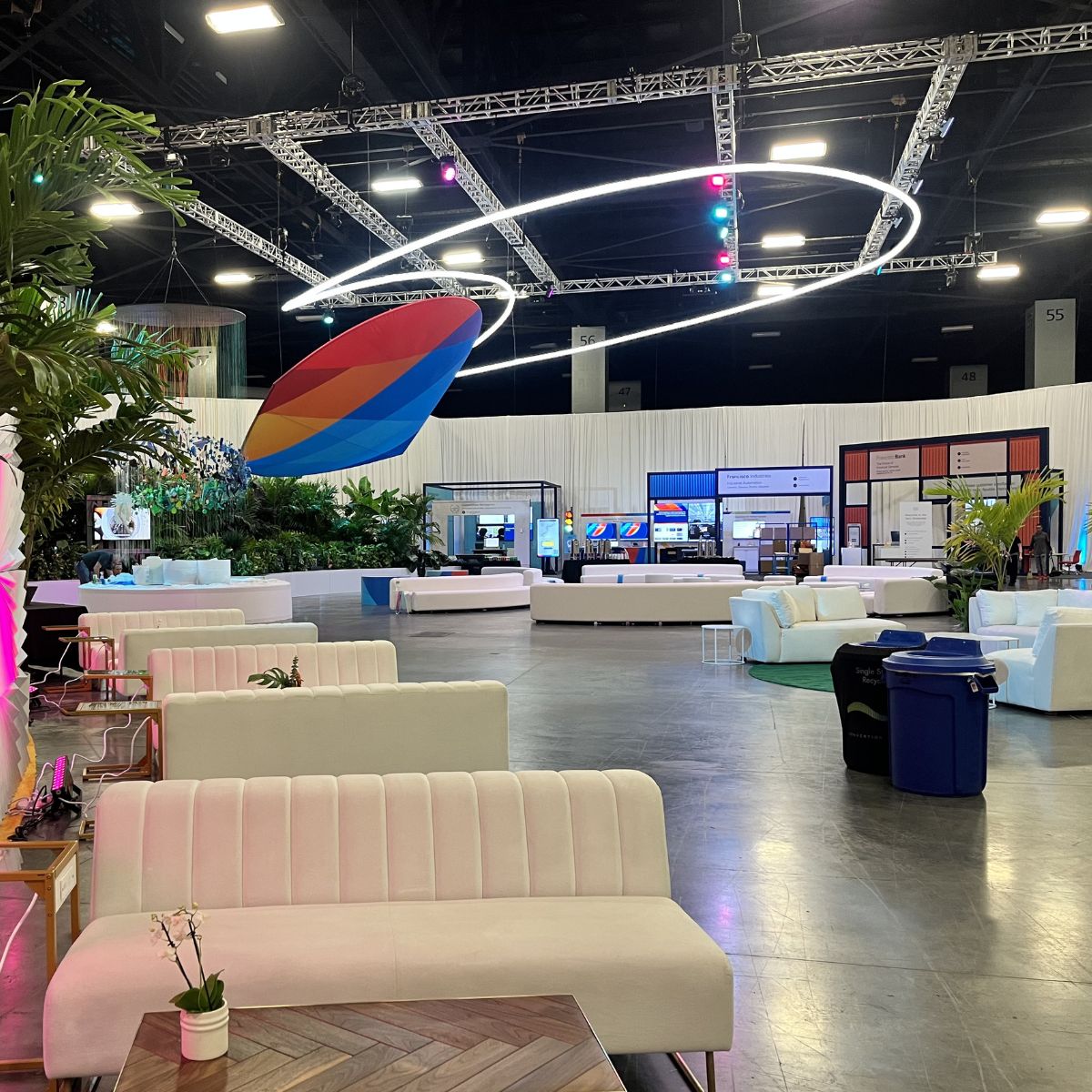 A spacious, modern event lounge features white sofas, potted plants, digital screens, and colorful lighting using the 4 Acrylic Tube for LED Lights ~ Model Smokies100. A large abstract hanging decoration adds to the inviting, organized feel.