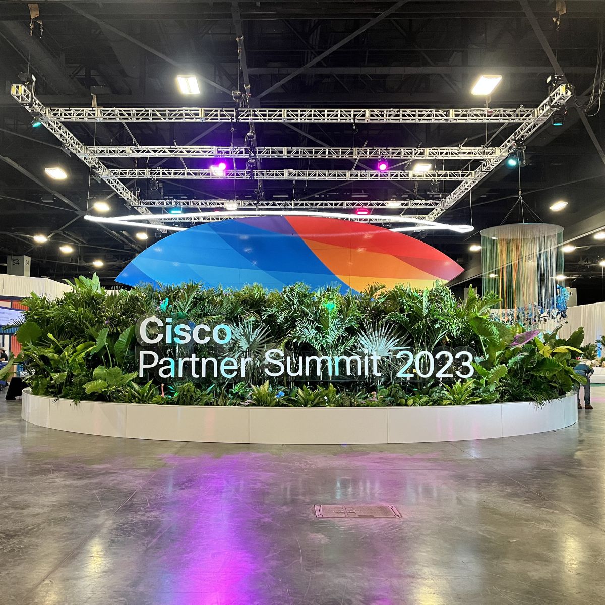 Large circular display with lush green plants and a sign reading Cisco Partner Summit 2023, indoors with colorful abstract shapes and Smokies100 4 acrylic tubes for LED lights integrated into the overhead lighting rigs.