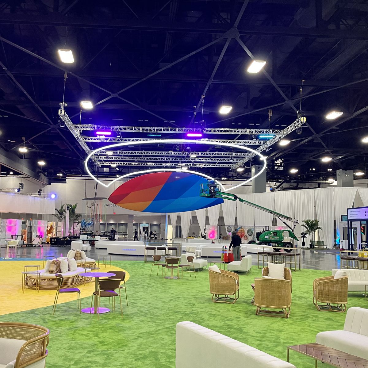 A large indoor event space features colorful seating, wicker furniture, and a high ceiling. In the center, a multicolored propeller hangs above a green lift and a Smokies100 4 Acrylic Tube for LED Lights.