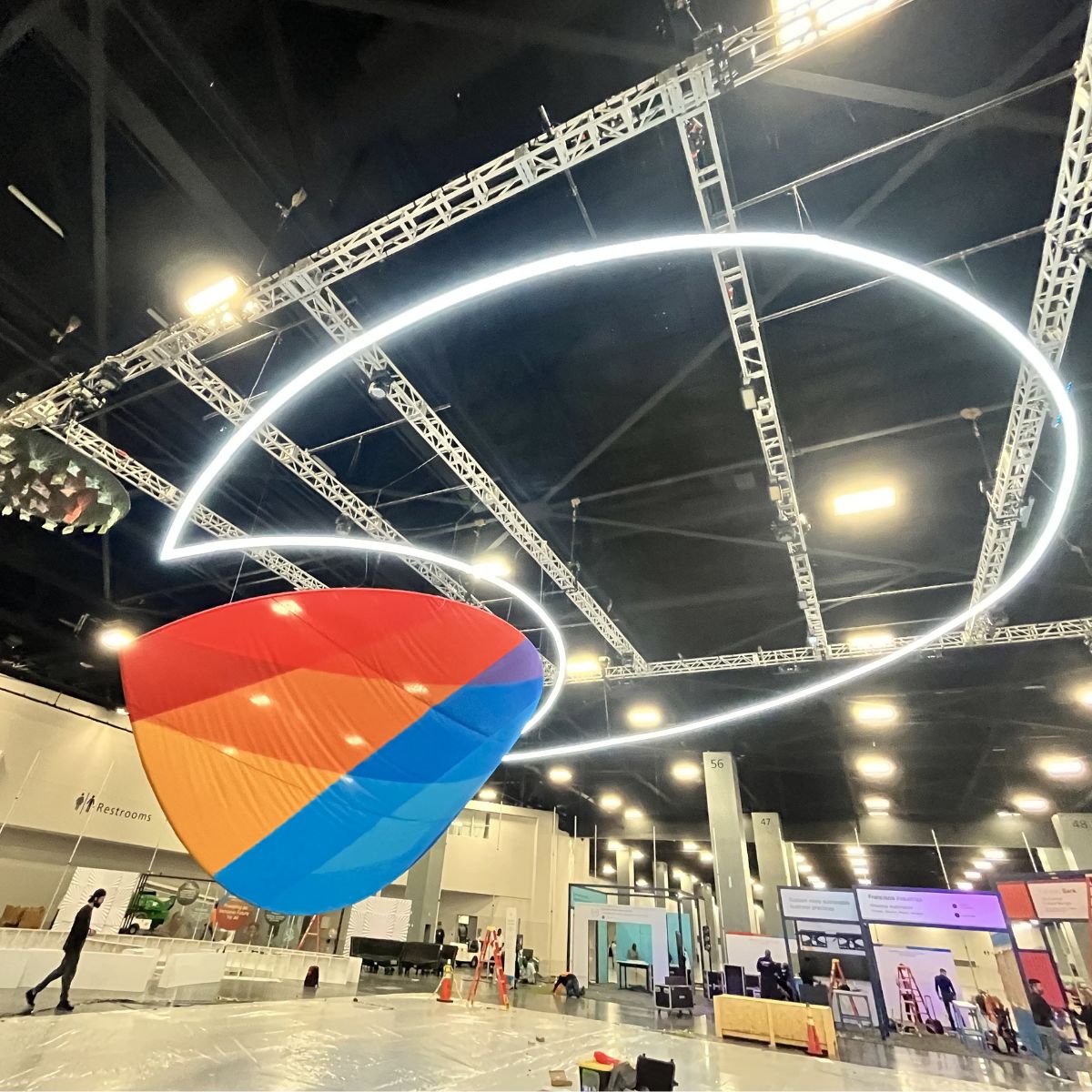 A vibrant geometric sculpture made from 4 Acrylic Tube for LED Lights (Model Smokies100) hangs from the event space ceiling, illuminated by bright lights and metal trusses as people set up displays and equipment below.