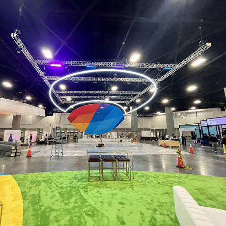 A large, colorful, oval balloon floats indoors under bright lights and circular trusses, with Smokies100 4 Acrylic Tubes for LED Lights illuminating green carpeted floors as people set up booths in the spacious convention center.