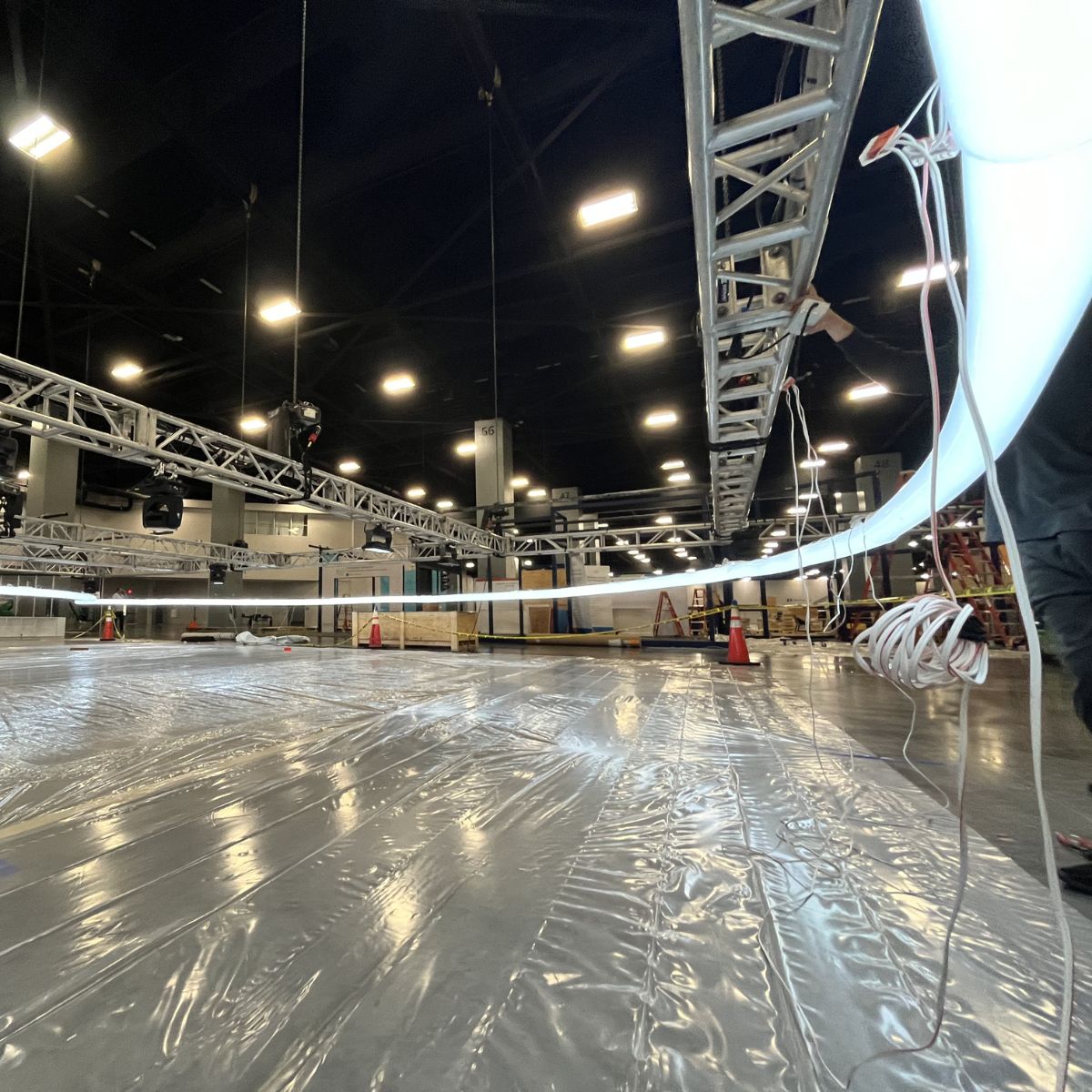 A large indoor venue with overhead lights, metal trusses, and cables. Workers are setting up equipment using 4 Acrylic Tube for LED Lights (Model Smokies100) with plastic sheeting on the floor for an event or production.