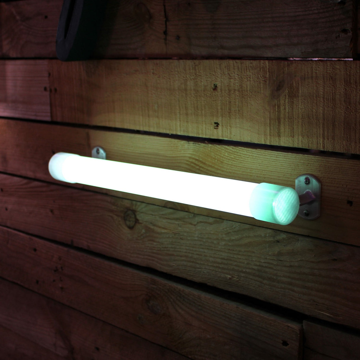 A Wired Post for 1.50 Round Profiles supports a glowing white LED diffuser tube, mounted horizontally on a wooden wall of long rectangular planks with visible mounting posts in brass or stainless steel for a modern look.