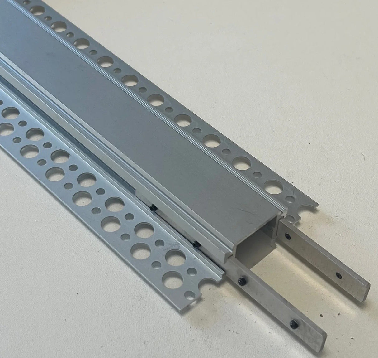 A close-up of a Flat Coupler Bracket for TL-041 Profile, featuring silver perforated flanges, a hollow rectangular channel, and two thin metal tabs extending from one end, resting on a light surface.