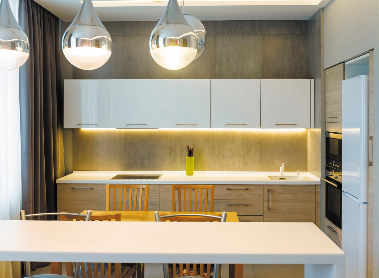 Modern kitchen features wooden cabinets, white countertops, and upper cabinets. Four wooden chairs line the bar with three metallic pendants. The 35/55 Degree Corner LED Lighting Channel (Model Texas Mini) adds a sleek accent near the stove and sink.