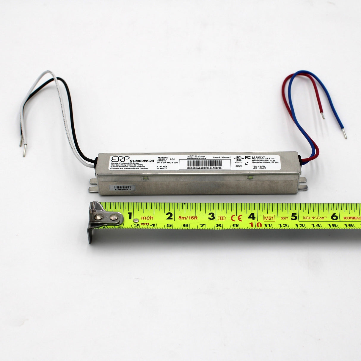 A Slimline Small Constant Voltage LED Driver (24V) from the ERP Power VLM Series, with wires at both ends, is placed above a yellow measuring tape showing its length of about 6 inches (15 cm); specs and constant voltage are shown on its label.