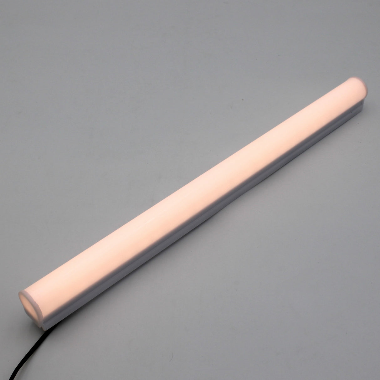 The White Border Contour Tube Light ~ Hi-Lite 30 Straight Section, a glowing cylindrical LED tube with a black power cord, lies diagonally on a plain light gray surface, ideal for building illumination.