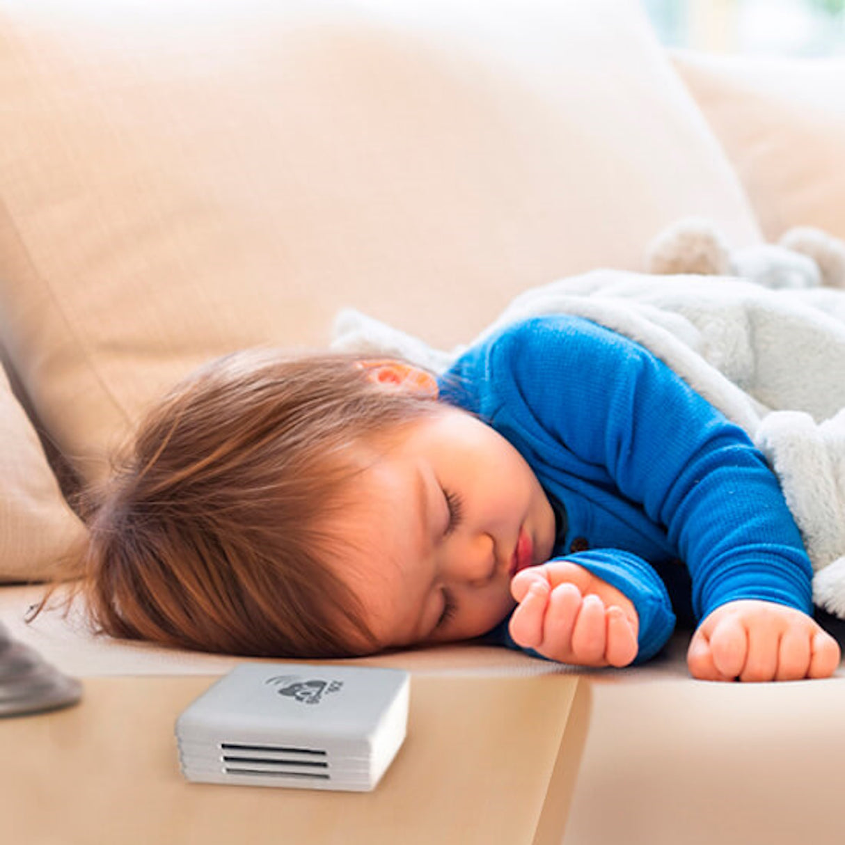 A toddler in a blue outfit sleeps on a beige couch, wrapped in a light blanket. The airSensor by Blebox—an Air Pollution Detector and Wi-Fi Air Quality Sensor—rests with ventilation slots and a warning symbol near the child’s hand.