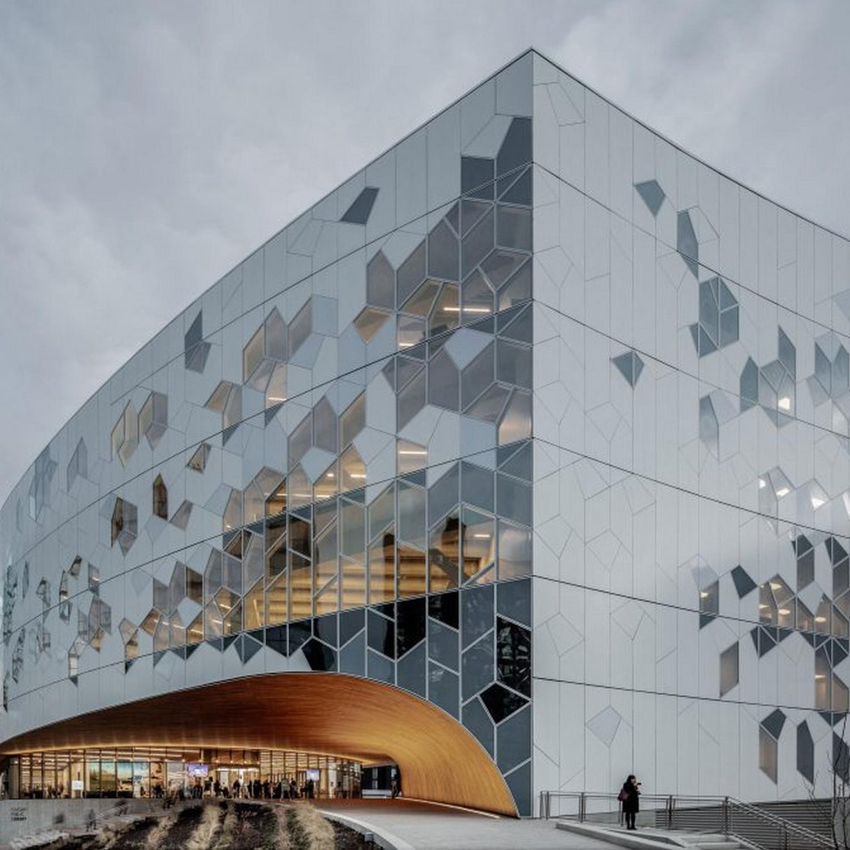 A modern building with a white geometric facade features hexagonal windows and Fire-Resistant Aluminum Composite Material Panel ~ ALPOLIC/fr Series. Large glass areas and a wood arch welcome people by the entrance on a cloudy day.