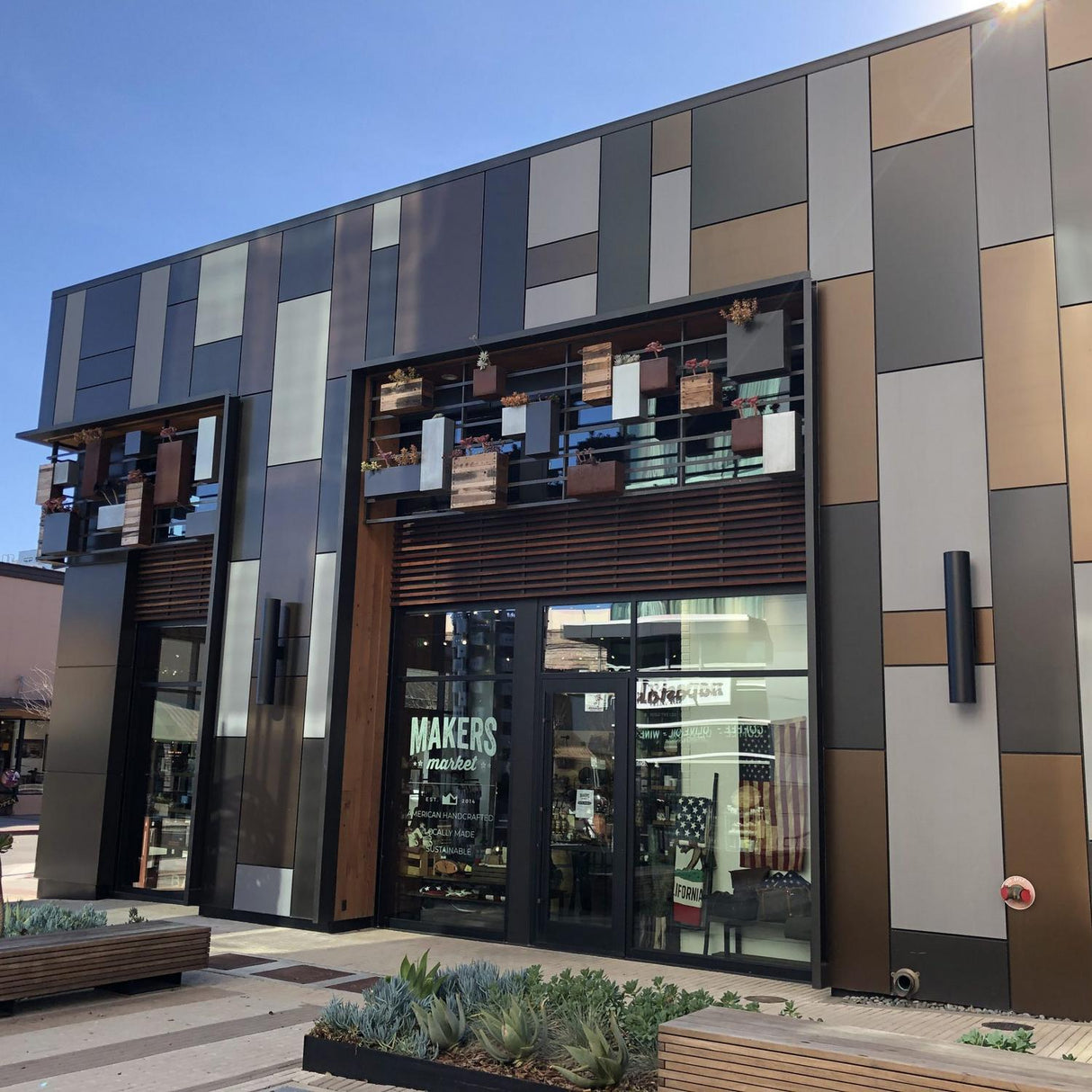 A modern building with a geometric metallic and neutral facade features large windows labeled MAKERS. The exterior uses ALPOLIC/fr Series fire-resistant aluminum panels. Planters with succulents accent the entrance and upper ledges.