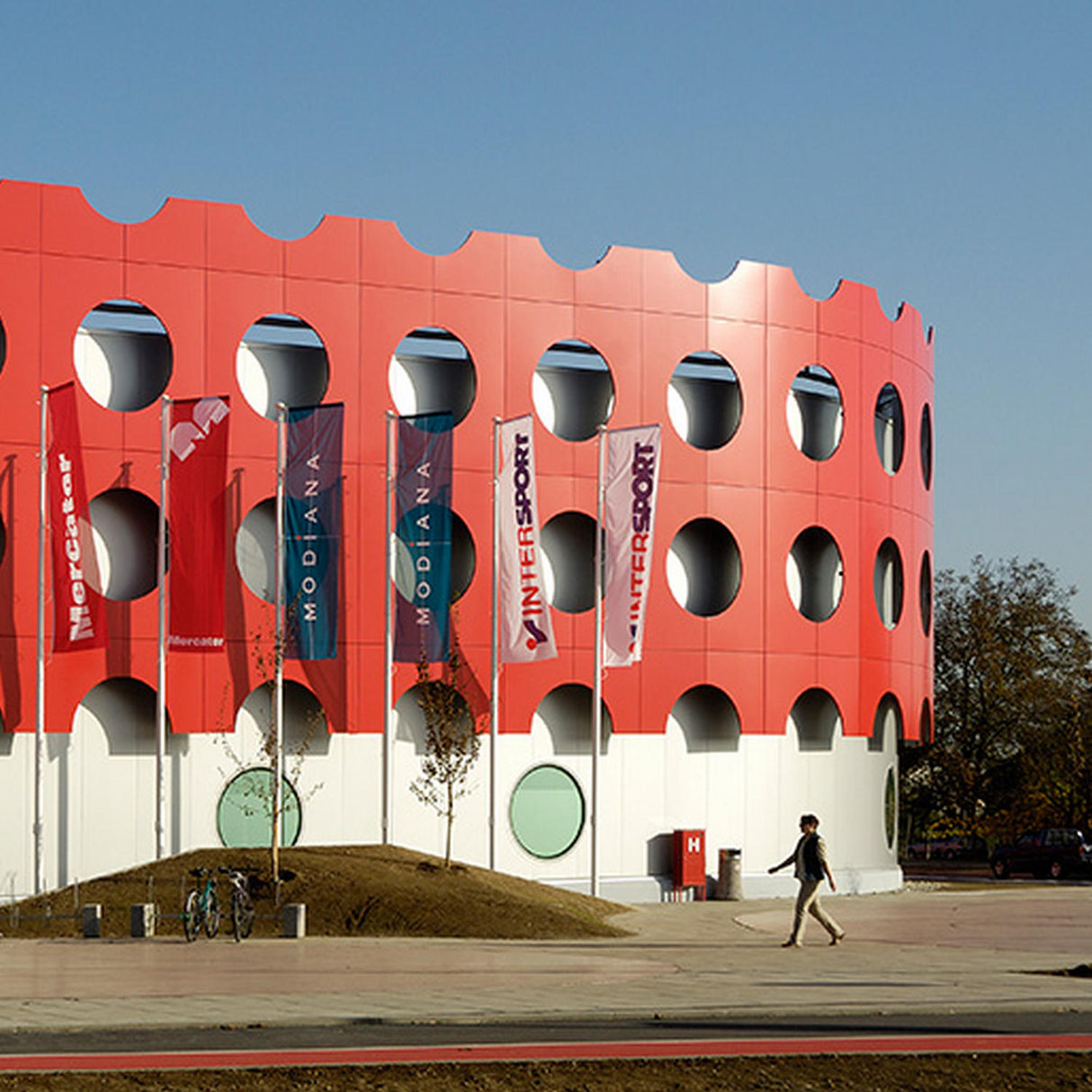 A person walks past a modern, circular building with a red and white facade featuring large round cutouts made from Heavy Duty Aluminum Composite Material Panel ~ ALPOLIC/HD Series. Store-name flags are displayed outside on a sunny day.