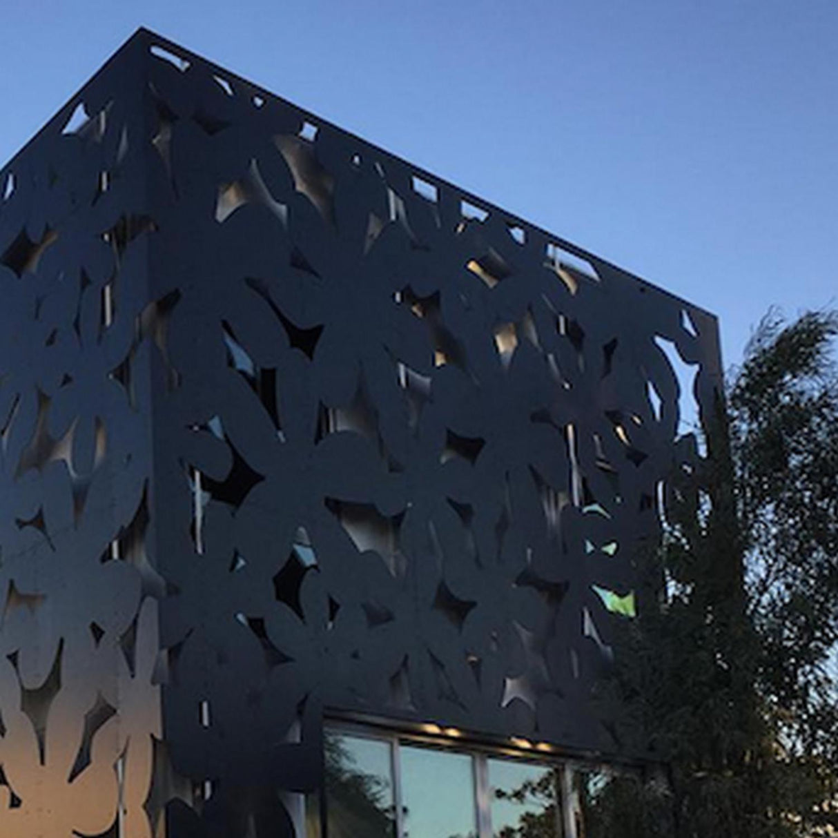 A modern building features a dark, leaf-patterned facade made from Heavy Duty Aluminum Composite Material Panel ~ ALPOLIC/HD Series, photographed at dusk against a clear sky, with trees visible on the right side.