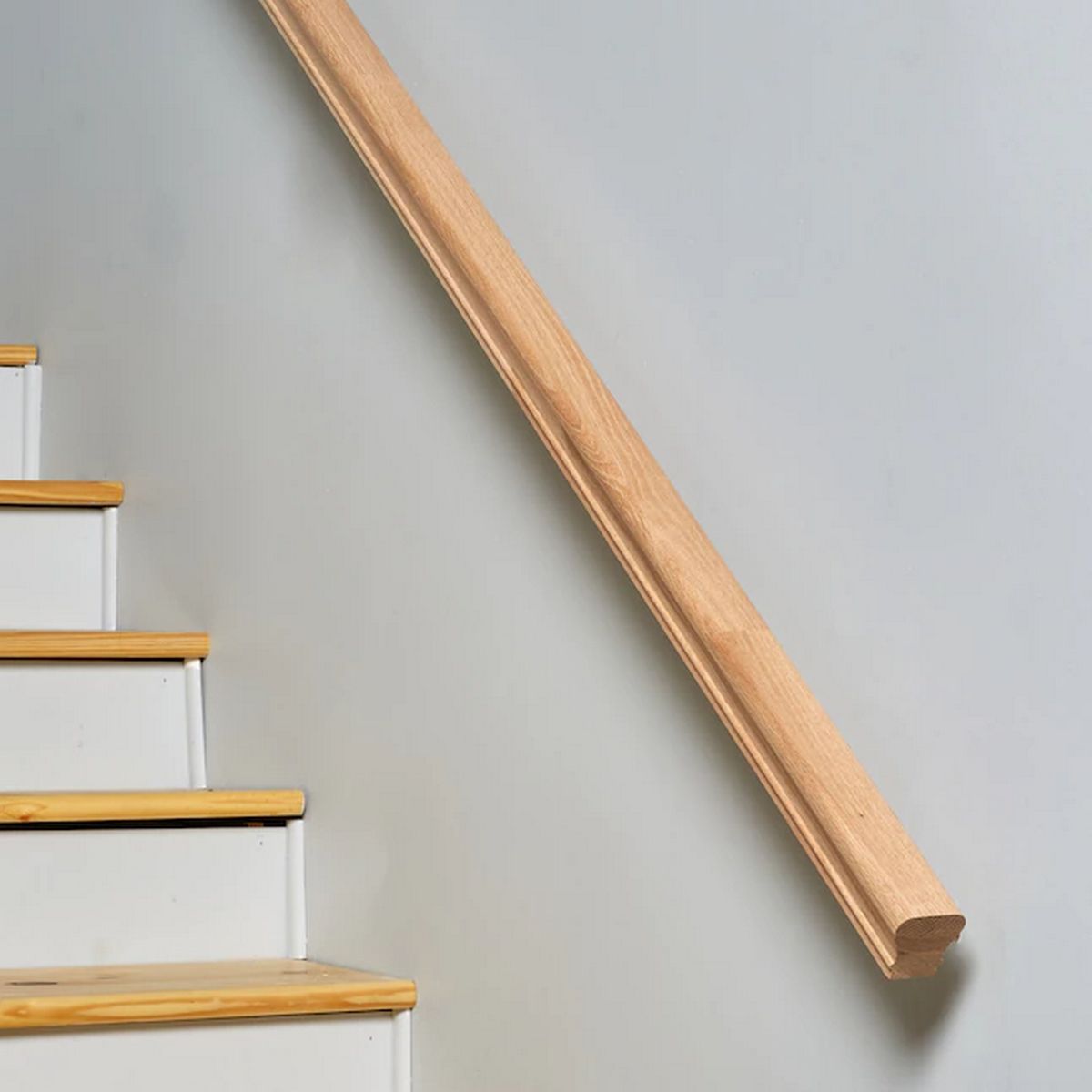 A Solid Wood LED Handrail Profile ~ Model Appalachians Wood is mounted diagonally on a white wall beside a staircase with white steps and light wooden treads.