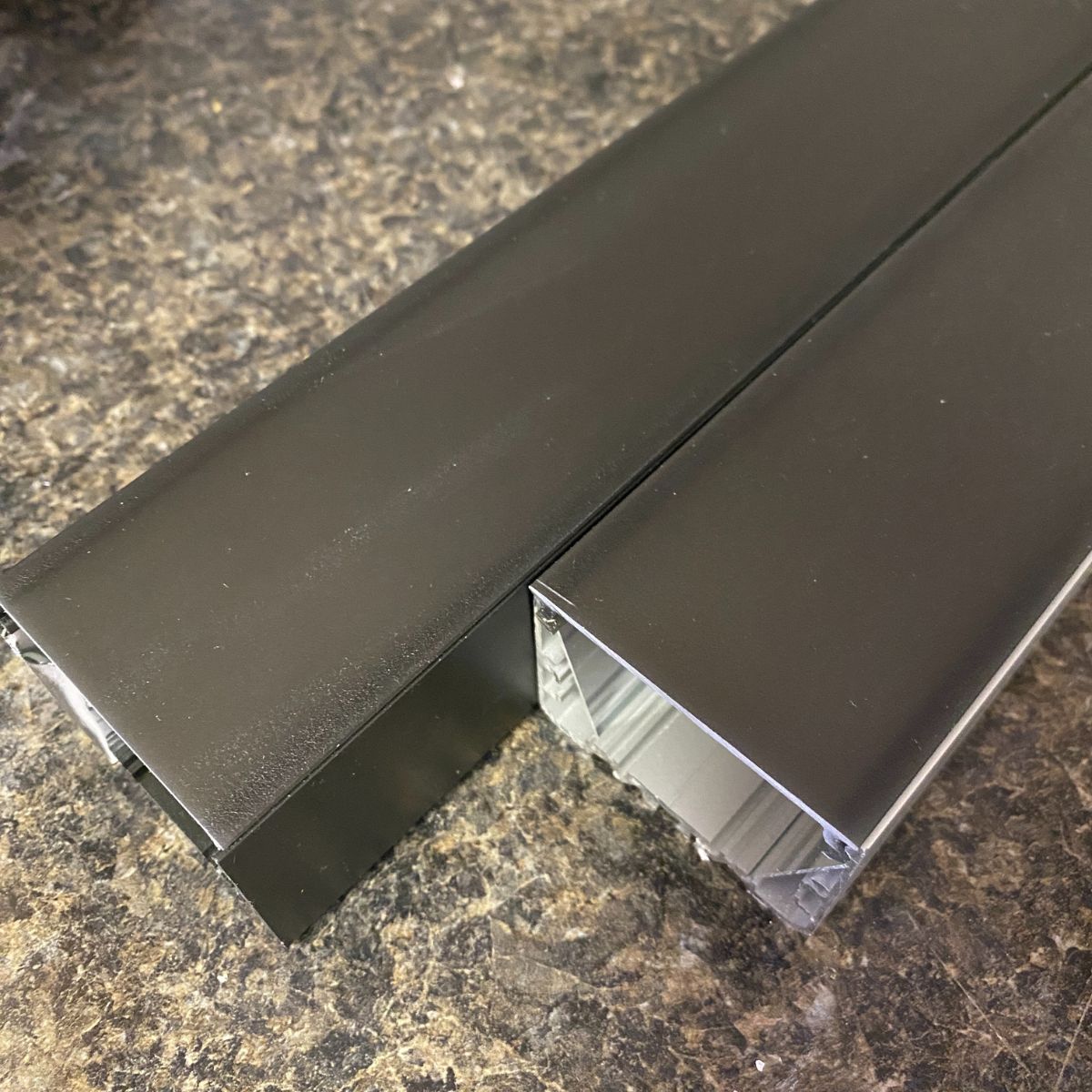 Two black rectangular Black Diffusers for PL55, DPL55, RPL55, and PL55-FL Profiles with hollow interiors rest on a speckled brown and black countertop—ideal LED profiles for effective light dispersion.