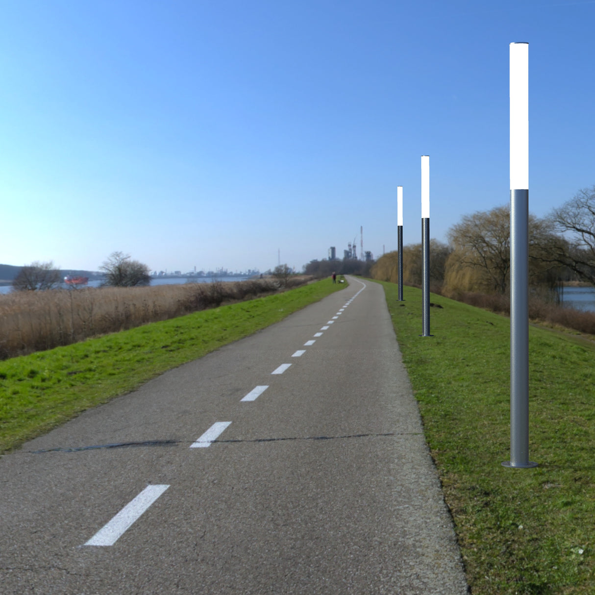 A paved riverside path bordered by grass and leafless trees features LED Bollard Post lights along the right side, with dashed white lines on the path and an industrial area visible in the distance.