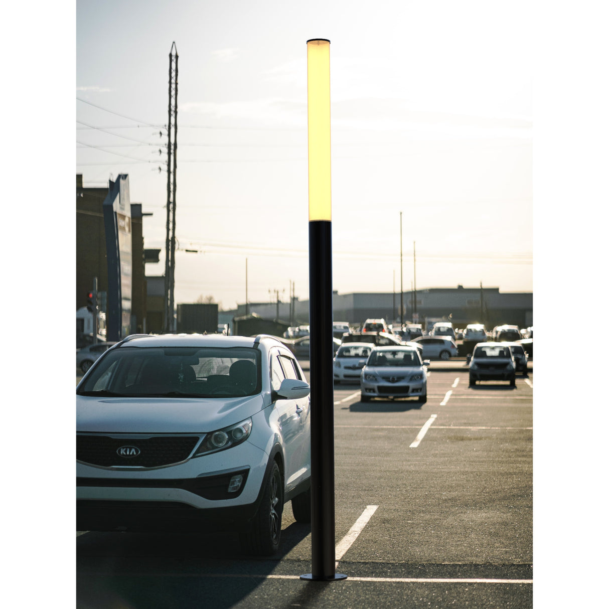 An LED Bollard Post with advanced lighting control stands in a parking lot at sunset, surrounded by parked cars and buildings in the background.