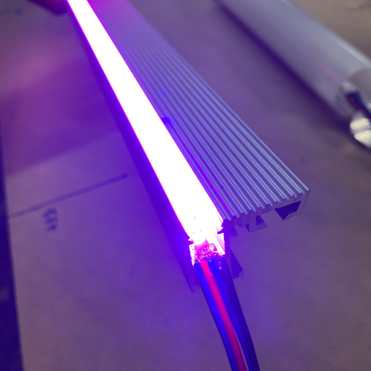 A close-up of the Cosmos Series Narrow IP20 RGB LED Strip (12V) glowing purple, mounted in a metallic grooved channel with exposed end wiring, set against a softly blurred neutral background.