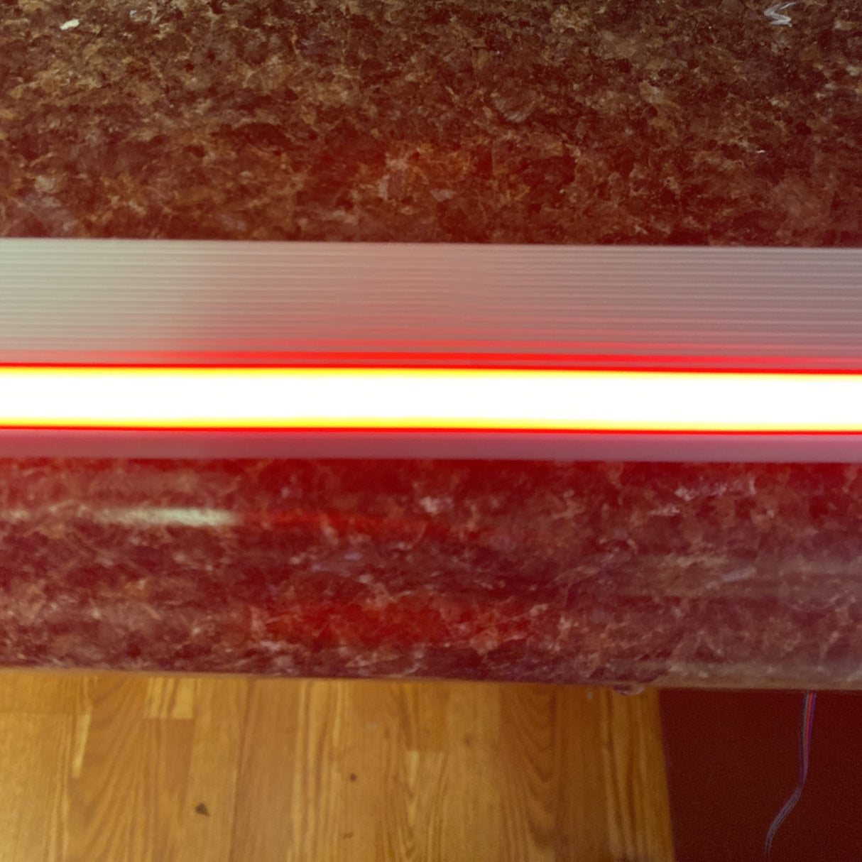 A close-up of a brown marble countertop features the Cosmos Series Narrow IP20 RGB LED Strip (24V), casting a glowing red light, with wooden flooring visible at the bottom.