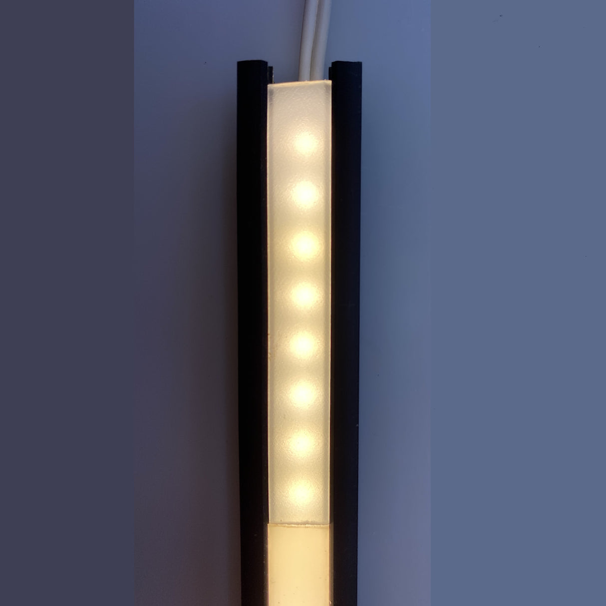 A vertical row of eight glowing LEDs is housed in the sleek black and white 0.39 Recessed LED Channel ~ Model Smart-In10, mounted on a light wall. Two wires extend from the top, creating a dot-free continuous line of illumination.