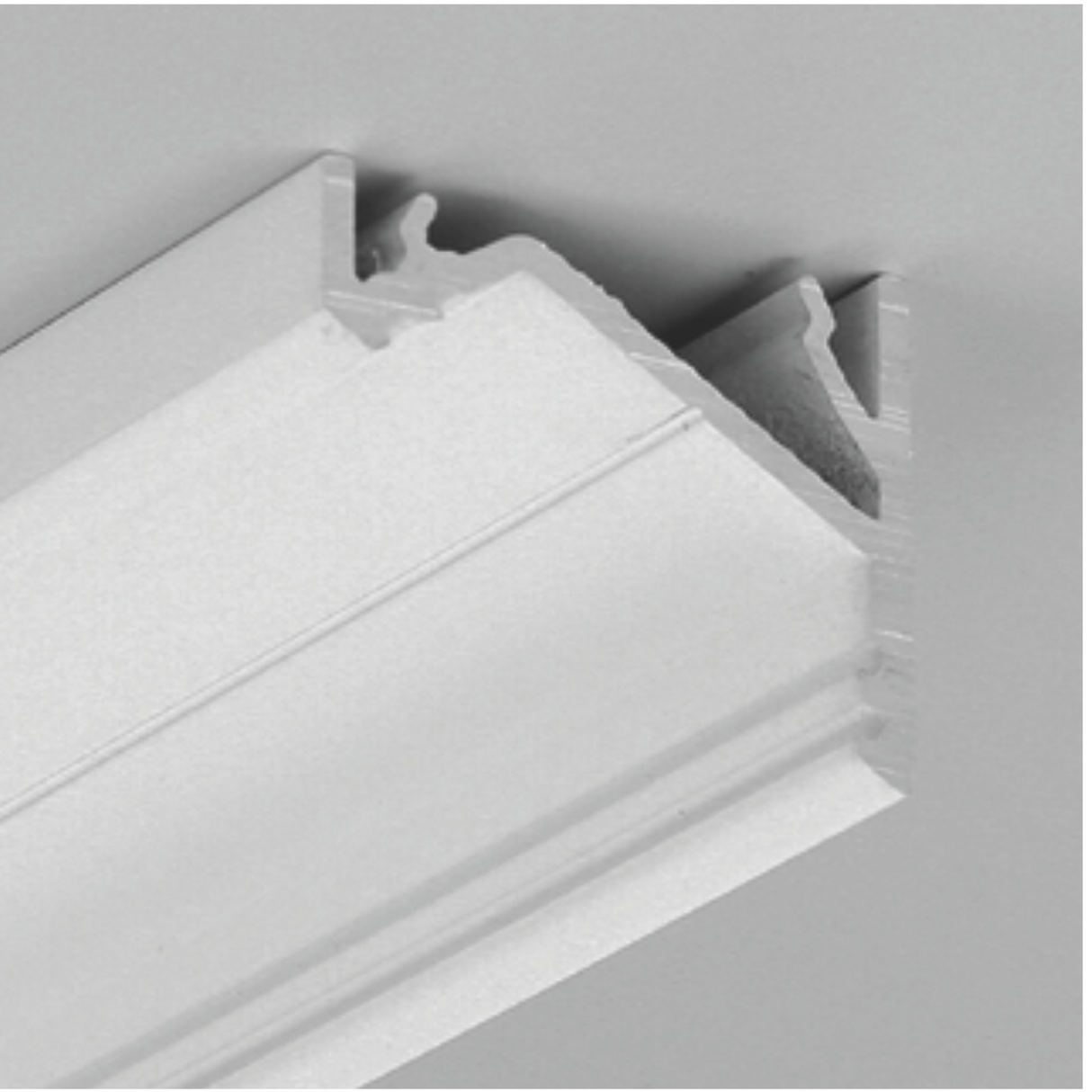 Close-up of the Surface Mount Corner LED Channel ~ Model Corner16, a white rectangular molding with a grooved, beveled profile, mounted at an angle—ideal for concealing under-cabinet lighting against a grey background.