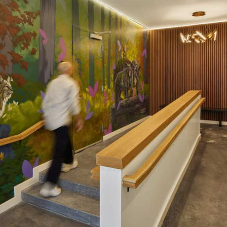 A person walks up steps beside a wooden handrail in a hallway with a vibrant forest mural, wood paneling, and subtle ceiling perimeter lighting from the Alu-Ceiling US Plaster-in Ceiling Perimeter Lighting Channel overhead.