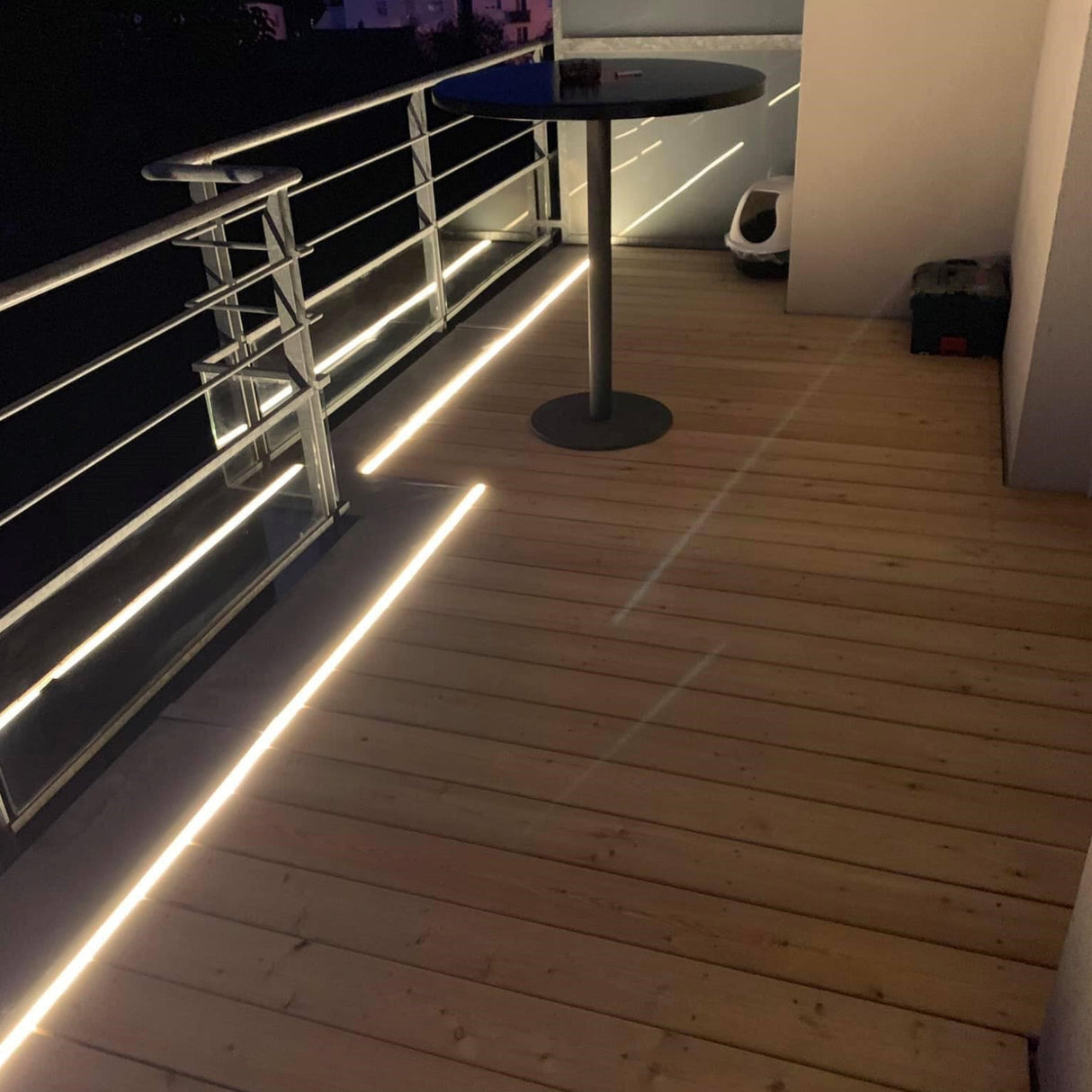 A balcony at night with wooden flooring features a Drive-safe Embedding LED Profile ~ Model Dublin XL along the railing, a round black table, a cat litter box, and a small black item in the corner.