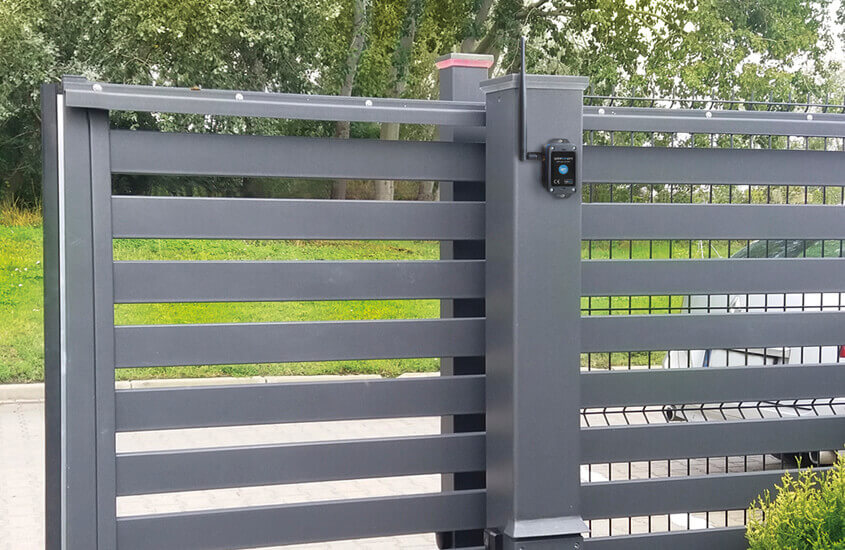 A modern black metal sliding gate with horizontal slats features the gateBox Pro by Blebox, an automatic gate and door controller for remote Wi-Fi operation, set outdoors amid green grass, trees, and paving.