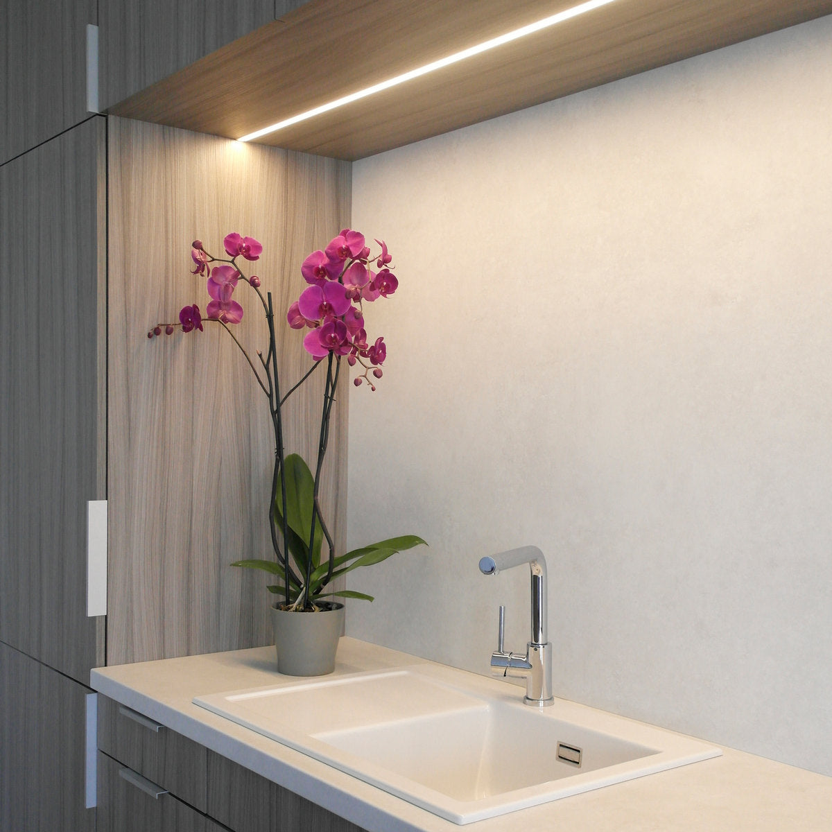 Modern kitchen countertop with a white sink, silver faucet, and potted purple orchid. Wooden cabinets and Groove10 0.39 Recessed LED Channels under the shelves brighten the minimalist space.