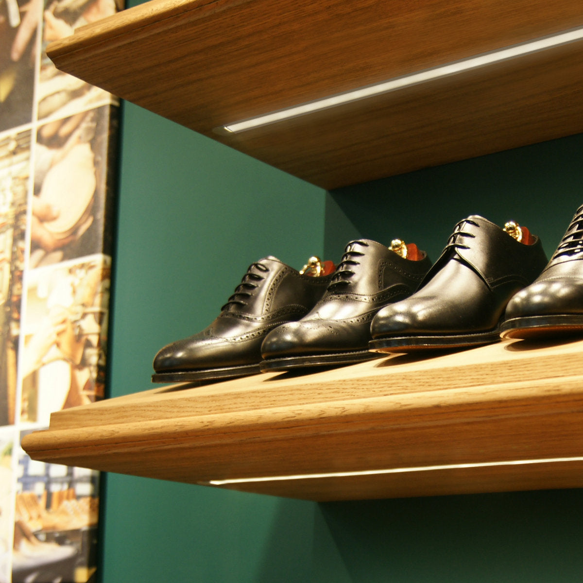 Three pairs of black leather dress shoes are displayed on wooden shelves against a green wall, subtly illuminated by 0.39 Recessed LED Channel (Model Groove10), with part of a shoe-making collage visible on the left side.