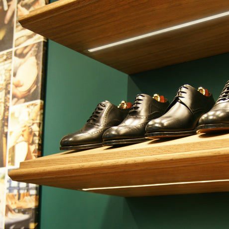 Three pairs of black leather dress shoes are displayed on wooden shelves against a green wall, subtly illuminated by 0.39 Recessed LED Channel (Model Groove10), with part of a shoe-making collage visible on the left side.