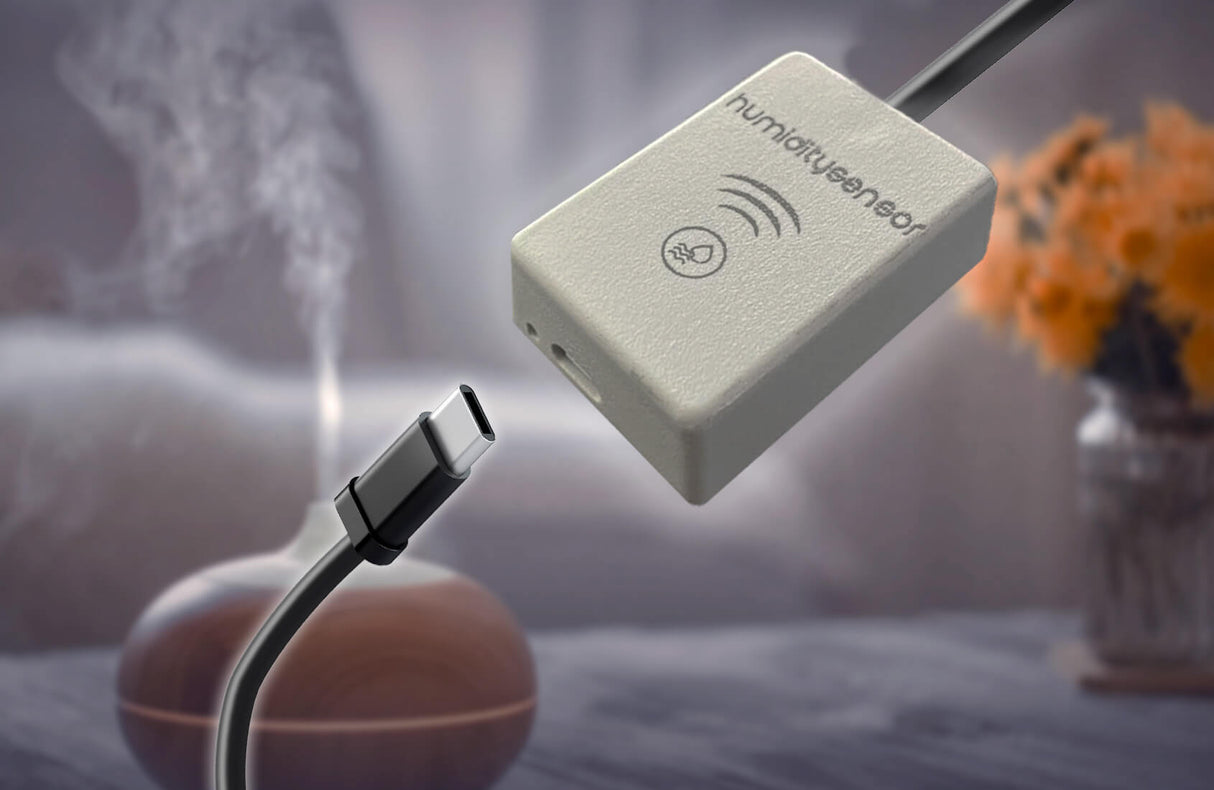 A USB-C cable sits next to a Smart Wi-Fi Air Temperature and Humidity Sensor ~ humiditySensor by Blebox. In the background, a humidifier mists near orange flowers, emphasizing smart climate monitoring.