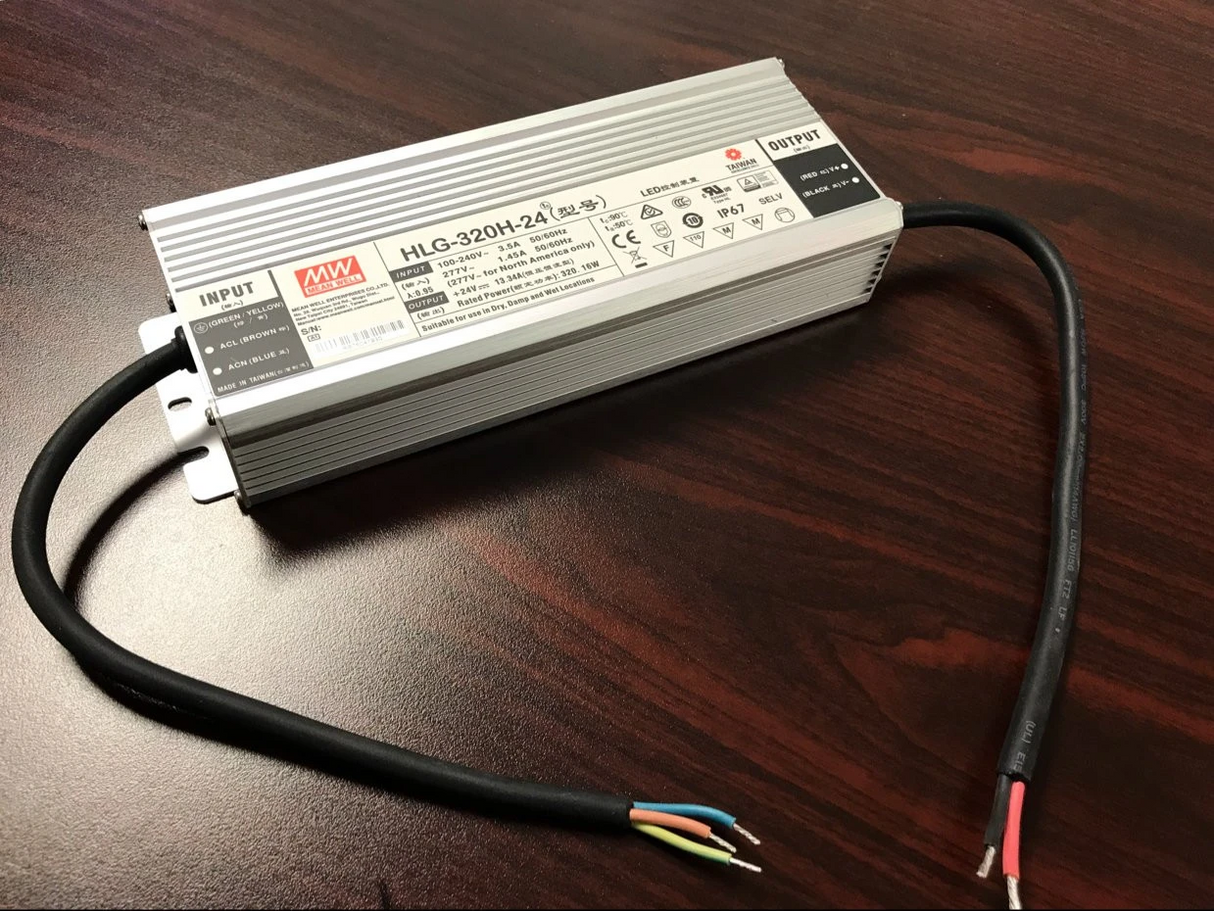 A silver Indoor / Outdoor LED Driver (24V) ~ Meanwell HLG Series with a black input cable and exposed wire ends rests on a dark wooden surface.