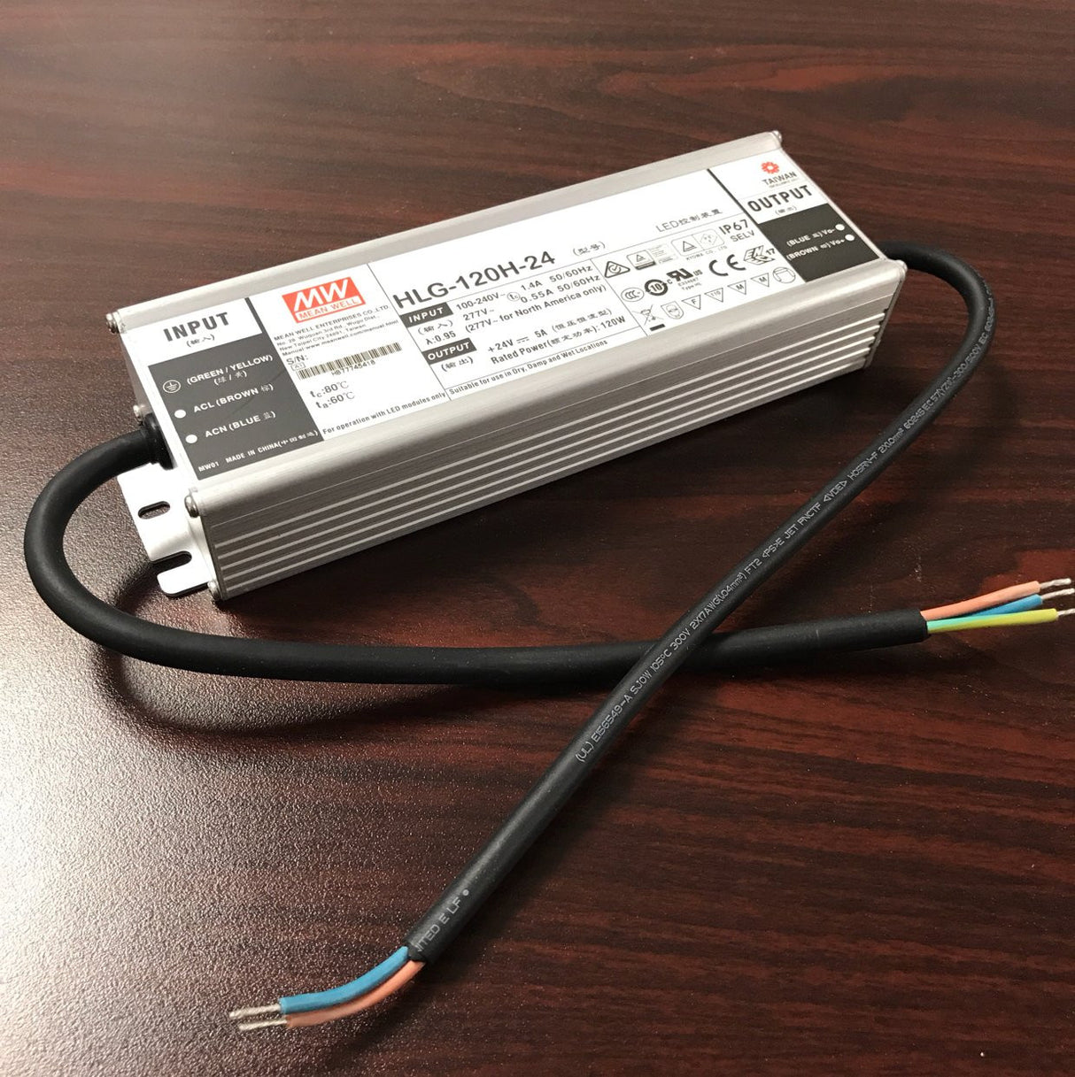 A silver Indoor / Outdoor LED Driver (24V) from the Meanwell HLG Series sits on a dark wood surface, with black input and output cables on each side and exposed colored wires at the end, illustrating its high efficiency.