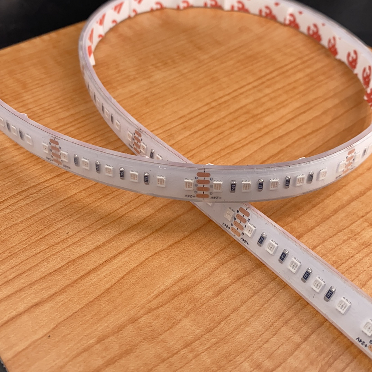 A close-up of two Bright RGB IP68 LED Strips (24V) ~ Verbena Series crossed on a light wood surface, showing their small LEDs and copper connectors.