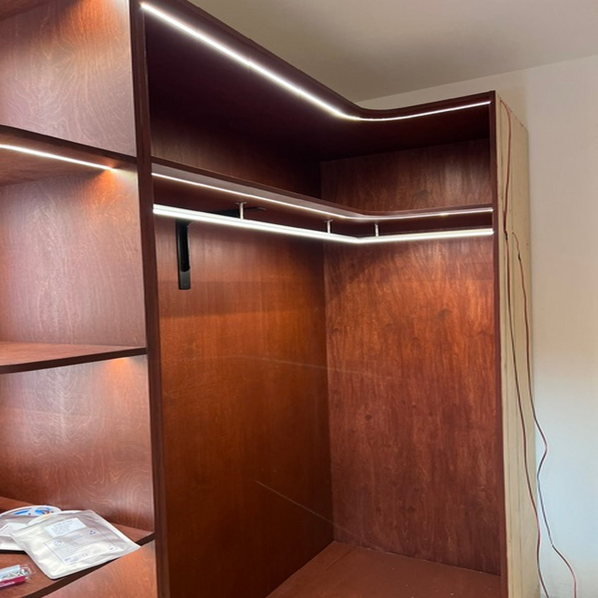 A wooden closet features the Lighted Closet Rod LED Channel ~ Model Alu-Round with an aluminum LED profile on shelf and top edges, a hanging rod supported by a black bracket, and open shelving on the left showing visible wires and packets.