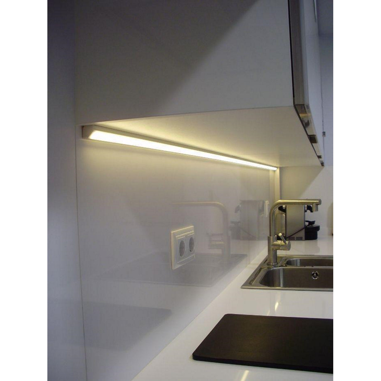 Modern kitchen countertop with a stainless steel sink, chrome faucet, and an ALU-45 45 Degree Corner Mount LED Channel lighting the glossy white backsplash. A double power outlet on the backsplash offers easy LED setup.